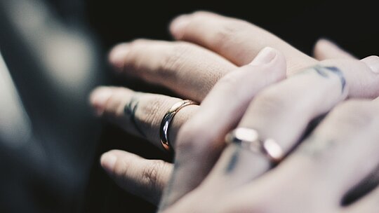 a foto mostra a mão de duas pessoas em close usando alianças de casamento e sobrepostas uma a outra