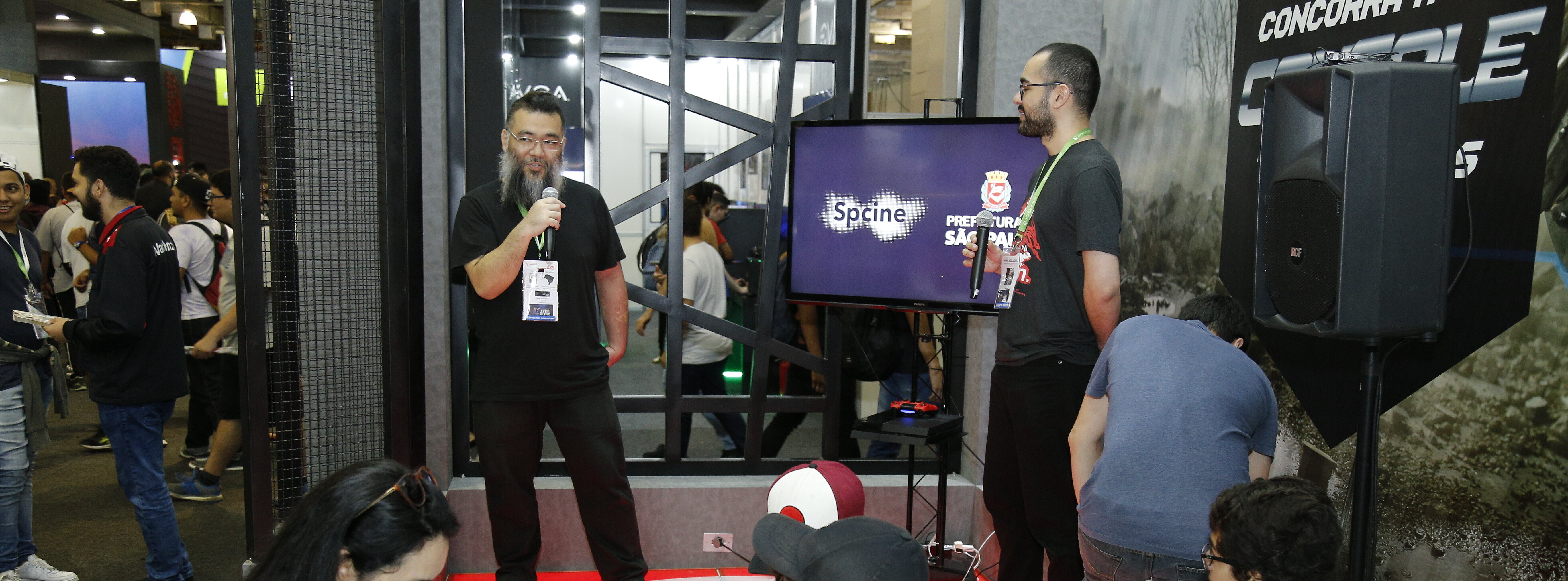 Dois palestrantes homens se apresentam sobre palco em estande no Mackenzie para plateia sentada e de pé.