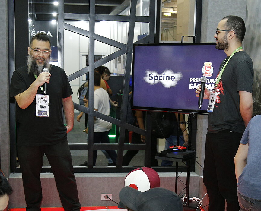 Dois palestrantes homens se apresentam sobre palco em estande no Mackenzie para plateia sentada e de pé.