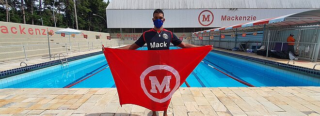Na imagem é apresentado o paratleta, Wendell Belarmino, perto da piscina segurando a bandeira do Mackenzie. 