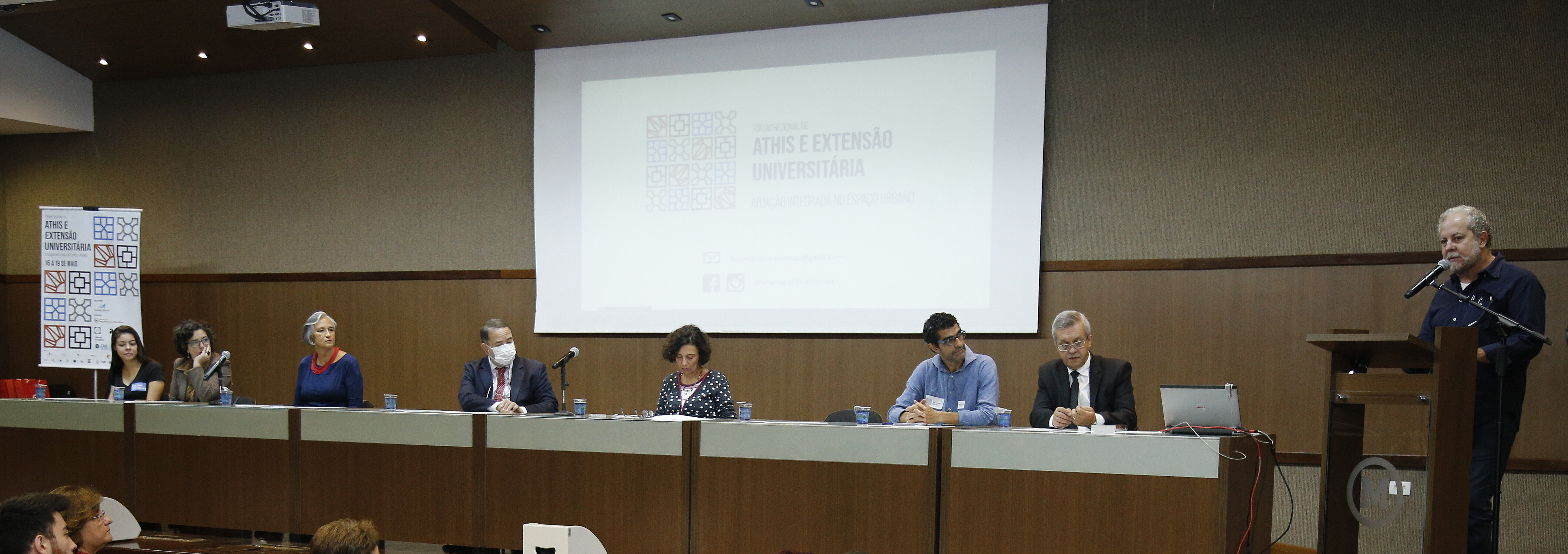 palestrantes sentados à mesa no palco e homem de pé ao púlpito