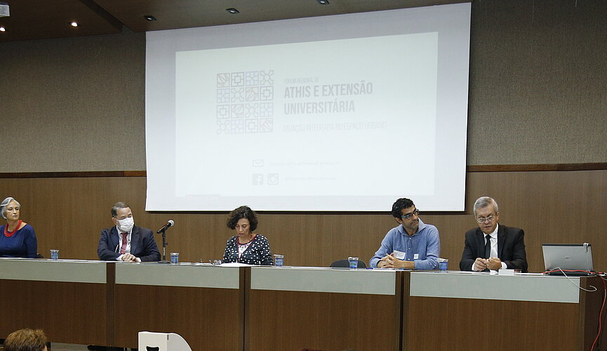 palestrantes sentados à mesa no palco e homem de pé ao púlpito