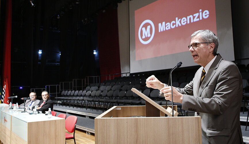 Simonaggio, ao púlpito, com telão ao fundo exibindo o logo do Mackenzie