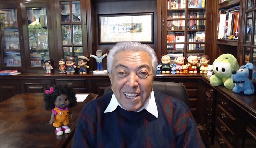 Maurício está em videoconferência com os alunos, em uma sala cheia de livros e personagens da Turma da Mônica