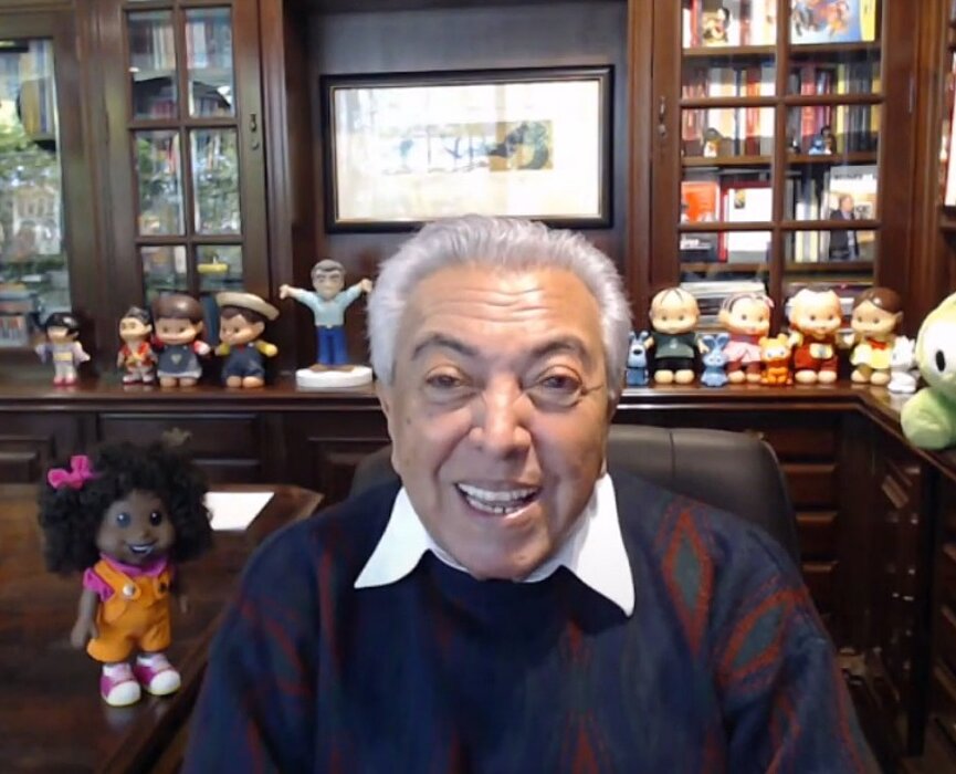 Maurício está em videoconferência com os alunos, em uma sala cheia de livros e personagens da Turma da Mônica