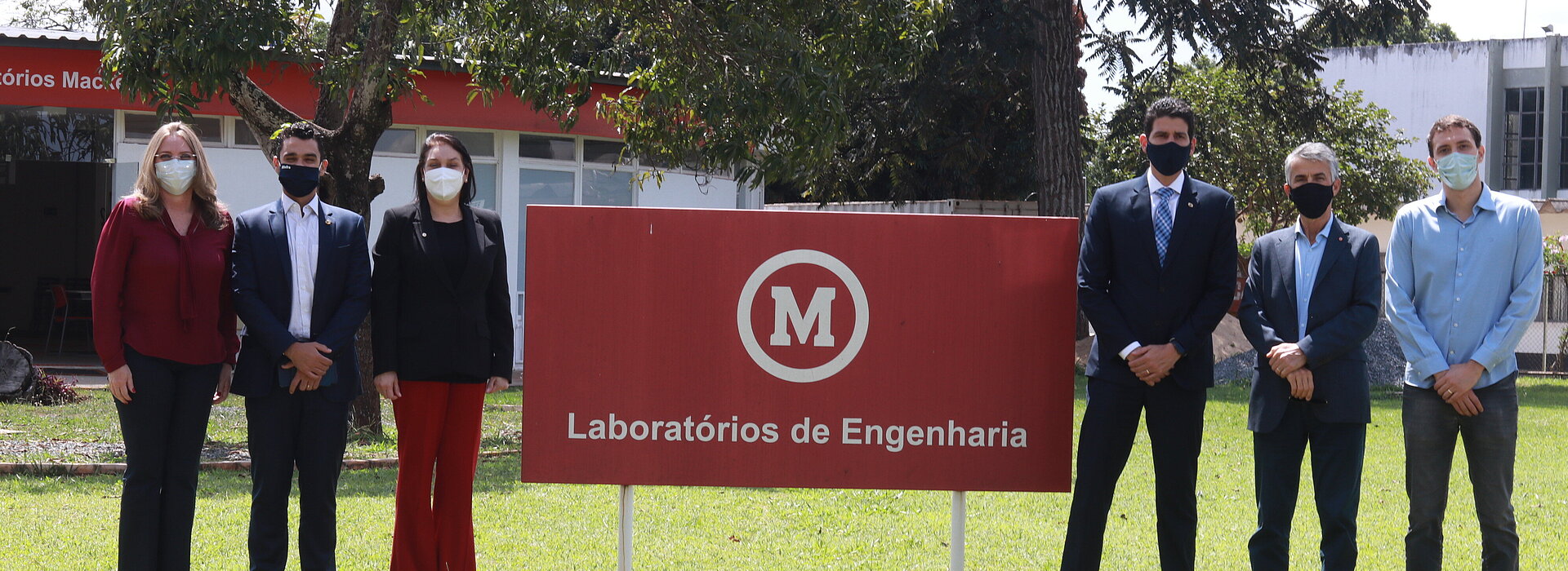 Seis pessoas, entre homens e mulheres, todos usando máscaras, posam para foto em um jardim de gramado verde ao lado da placa que diz, "Laboratório de Engenharia"