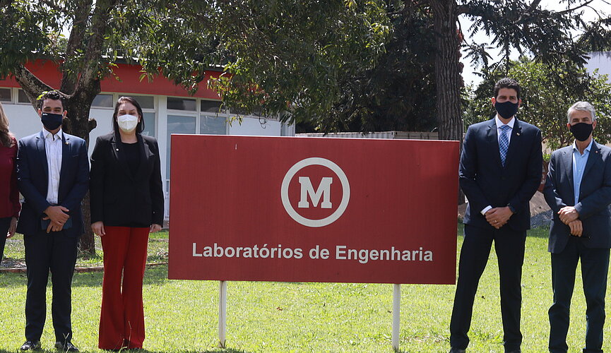 Seis pessoas, entre homens e mulheres, todos usando máscaras, posam para foto em um jardim de gramado verde ao lado da placa que diz, "Laboratório de Engenharia"