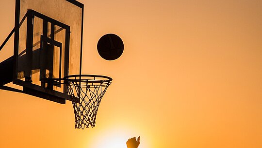 Imagem mostra uma cesta de basquete com um homem jogando a bola em direção a ela.