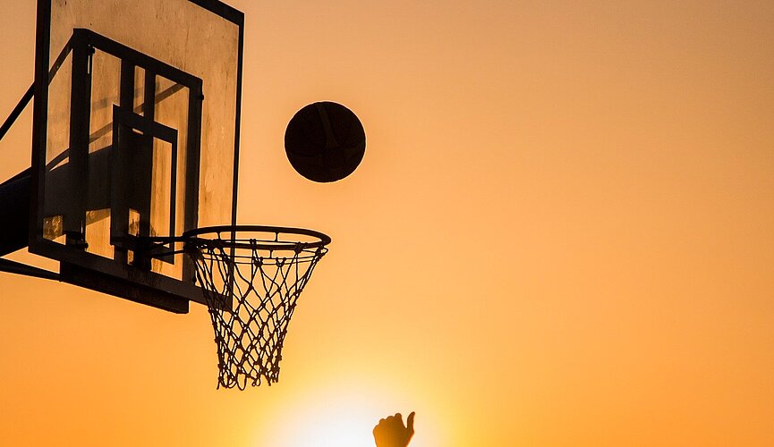 Imagem mostra uma cesta de basquete com um homem jogando a bola em direção a ela.