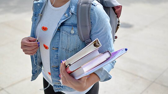 A imagem apresenta uma estudante carregando livros e cadernos. 