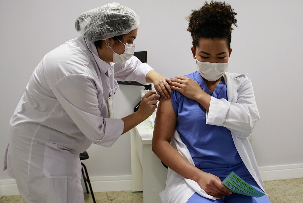 mulher sentada de camisa azul e jaleco branco recebe vacina no braço de outra mulher paramentada com luvas, touca, jaleco e afins. Ambas estão usando máscaras