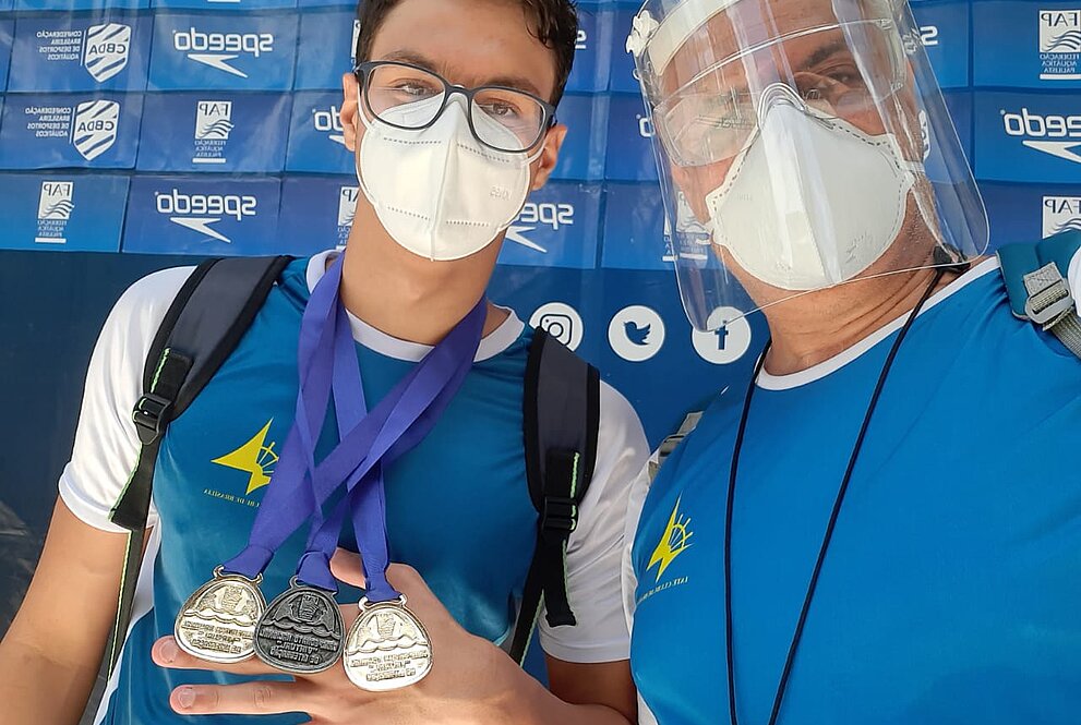 Rapaz com máscara exibe medalhas conquistadas em competições ao lado de um homem com proteções faciais