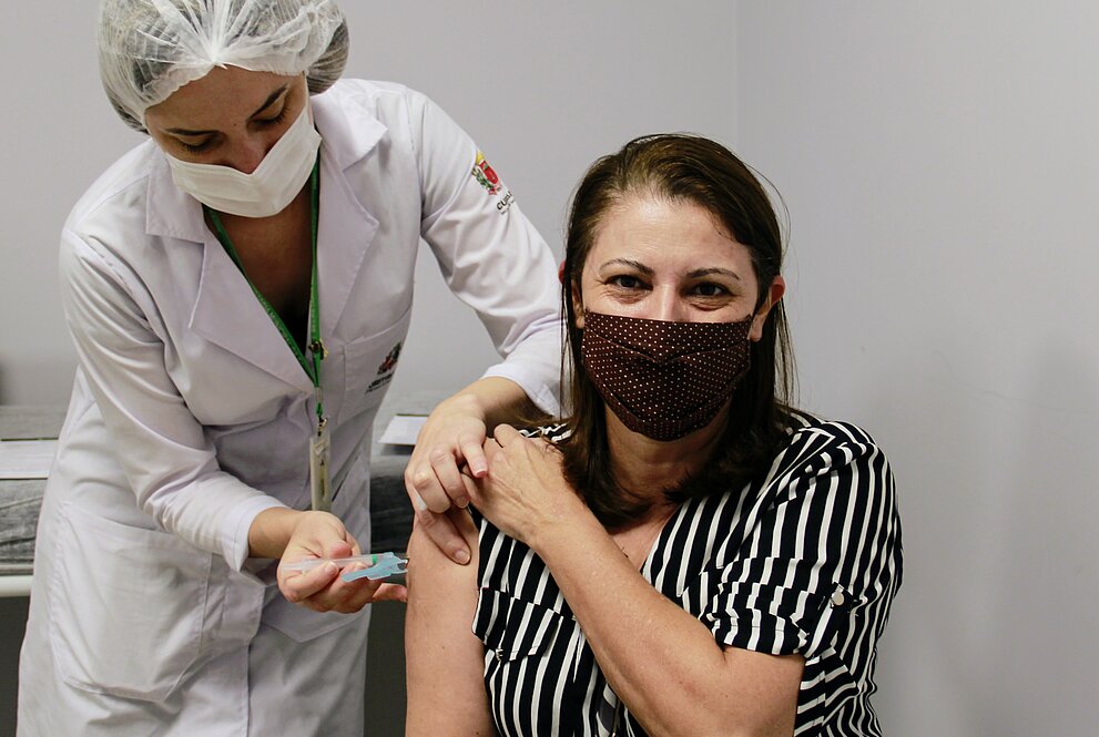mulher branca, sentada usando camisa listrada em tons preto e branco recebe vacina de mulher branca de pé usando jaleco, luvas e touca. Ambas usam máscaras.