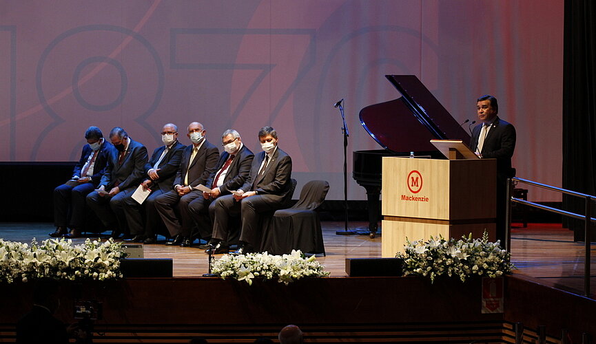 Palco com seis homens assentados enquanto outro fala em um púlpito