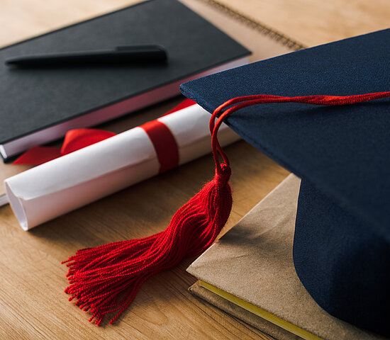 Imagem de um Chapéu de formatura, um caderno e um diploma em cima de uma mesa.
