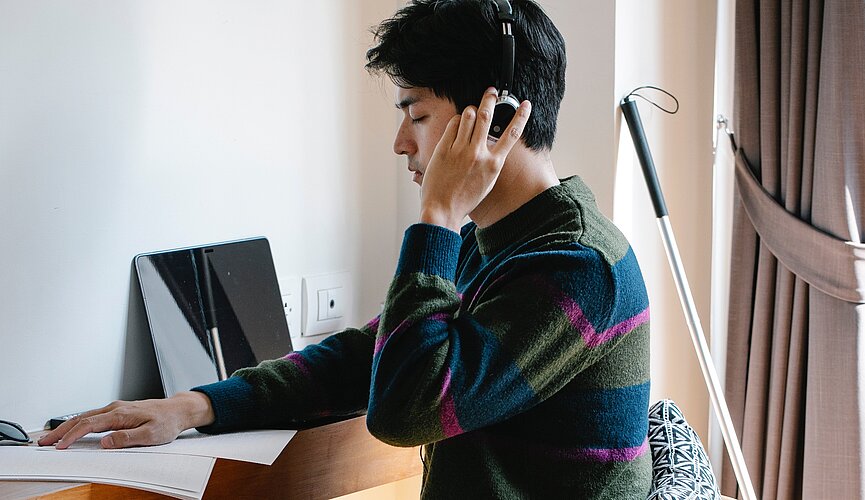 A foto mostra um homem sentado em frente a um computador fazendo a leitura em braile de um livro com sua bengala vermelha atrás