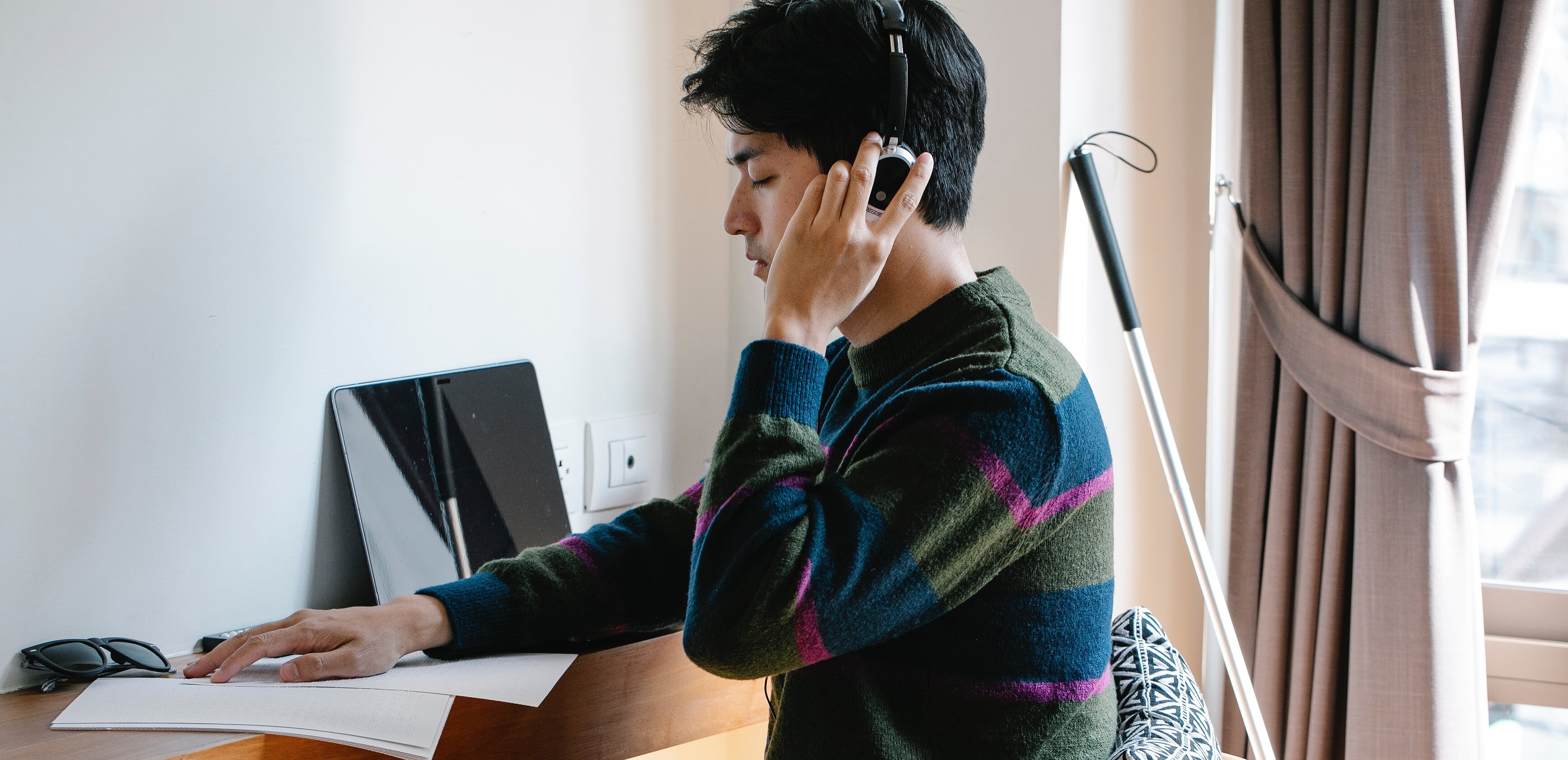 A foto mostra um homem sentado em frente a um computador fazendo a leitura em braile de um livro com sua bengala vermelha atrás