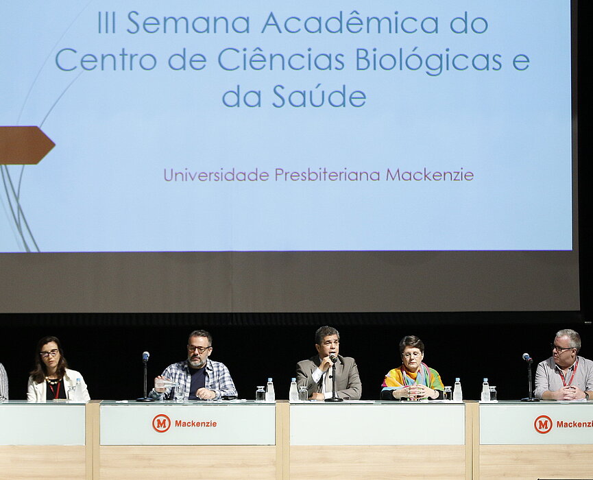 autoridades sentadas à mesa durante evento. Telão na parte de trás da imagem