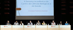 autoridades sentadas à mesa durante evento. Telão na parte de trás da imagem