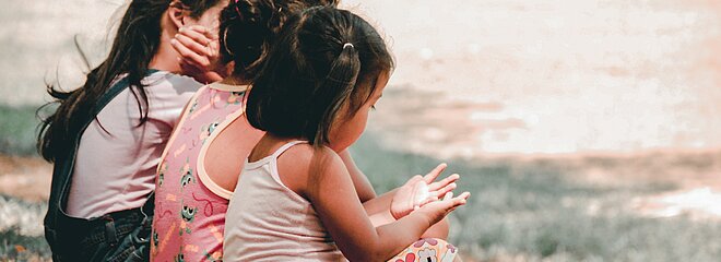 A foto mostra três crianças sentadas de costas em um campo de grama