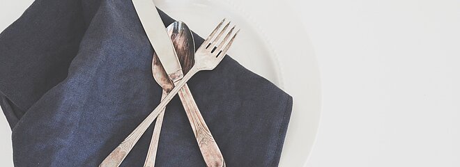 Imagem com um prato, um guardanapo preto de tecido em cima, uma faca, um garfo e uma colher