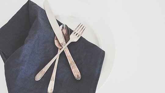 Imagem com um prato, um guardanapo preto de tecido em cima, uma faca, um garfo e uma colher