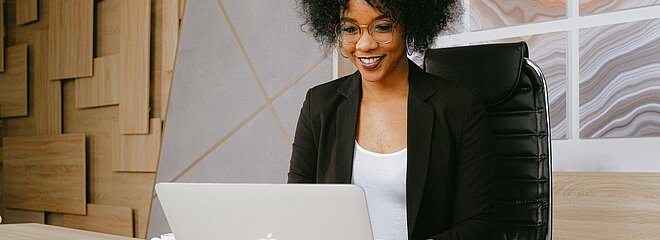 Uma mulher nova está sentada em uma cadeira em frente ao computador. Está sorrindo e digitando. 