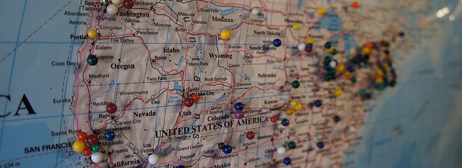 Foto mostra o mapa dos Estados Unidos com vários alfinetes redondos coloridos.  