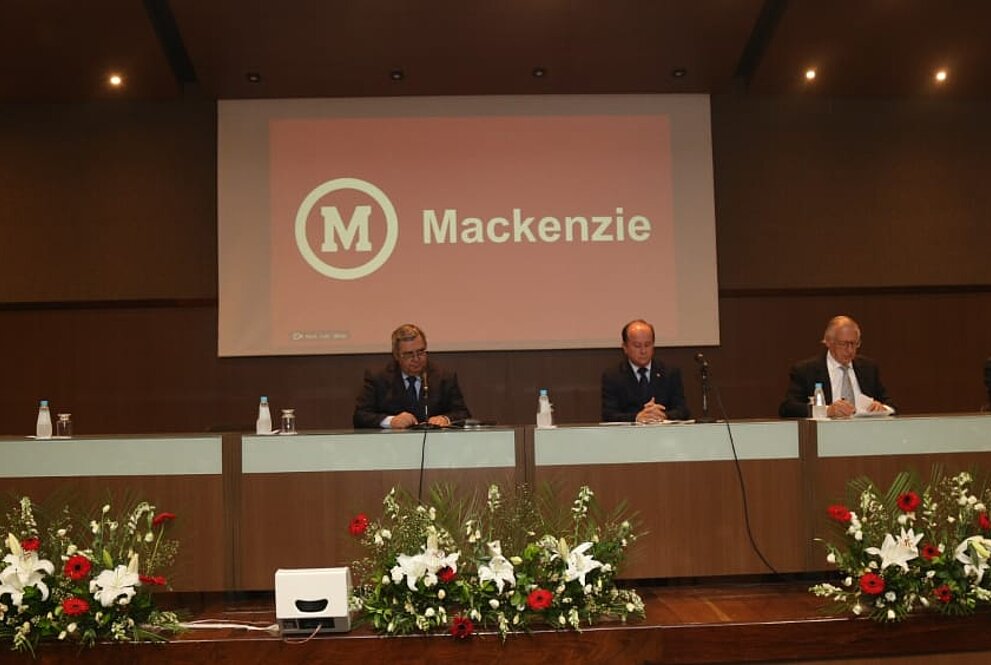 Autoridades mackenzistas sentadas à mesa do evento com tela projetando nome "Mackenzie" ao fundo