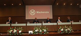 Autoridades mackenzistas sentadas à mesa do evento com tela projetando nome "Mackenzie" ao fundo