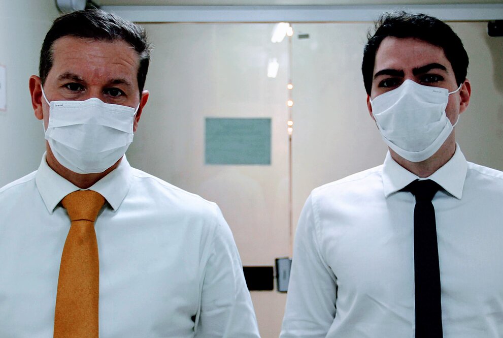 Dois médicos com camisa, gravata e máscara estão no corredor de um hospital com paredes claras