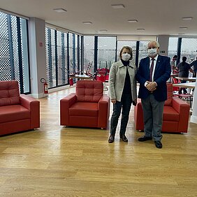 Na imagem, um homem e uma mulher estão de pé em uma sala com poltronas vermelhas e chão de madeira com grandes janelas em torno