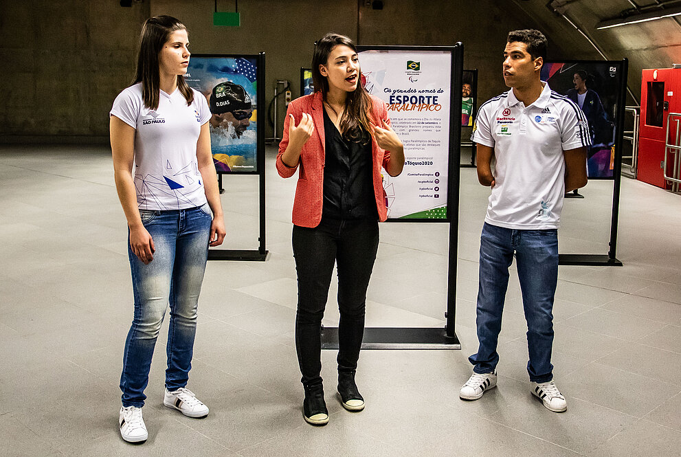 Alana Maldonado ao lado de Cinthia do Carmo e Daniel Dias em meio aos painéis da exposição