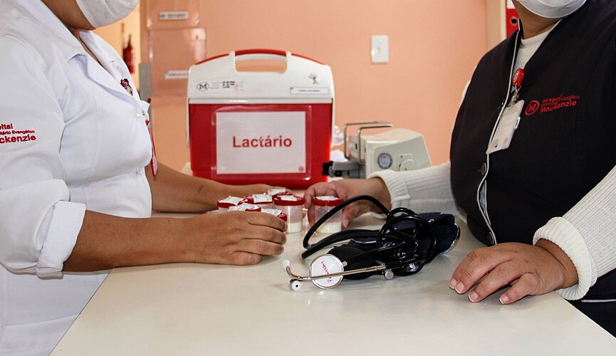 Na foto, as mãos de duas mulheres aparecem sobre um balcão branco onde estão apoiados um estetoscópio e uma caixa térmica vermelha