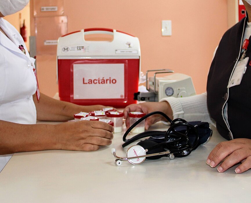 Na foto, as mãos de duas mulheres aparecem sobre um balcão branco onde estão apoiados um estetoscópio e uma caixa térmica vermelha