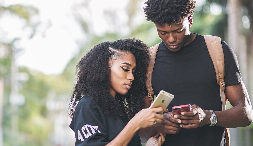 Na imagem, duas pessoas conferem um celular. Ao fundo, pode-se ver árvores e parte do céu