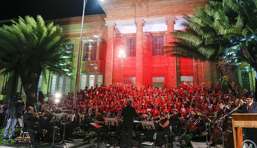 Imagem mostra coral do Mackenzie durante cantata de Natal realizada em 2018. A foto tem árvores, estrutura do seminário de tijolinhos, luz vermelha e o coral. 