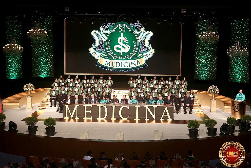 Turma de medicina no palco, sentados usando becas e com telão ao fundo com símbolo da medicina