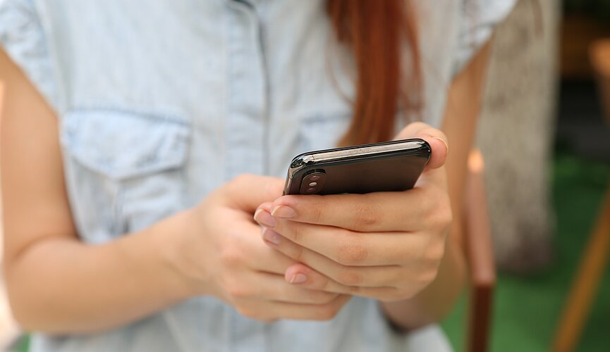 A foto mostra uma mulher com cabelo comprido mexendo no celular. Seu rosto não aparece, apenas seus braços e as mãos segurando o celular. 