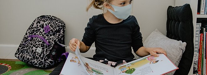 A foto mostra uma criança sentada no chão, com um livro aberto. Ao seu lado tem uma mochila. A criança está usando máscara contra a covid-19.