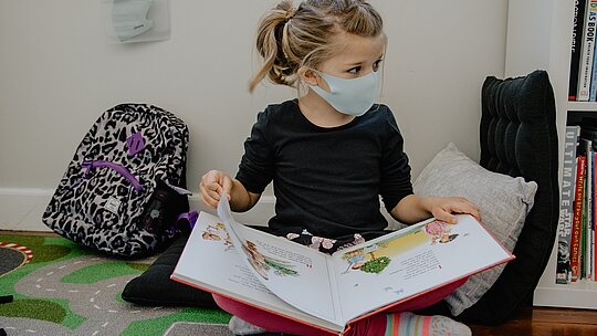 A foto mostra uma criança sentada no chão, com um livro aberto. Ao seu lado tem uma mochila. A criança está usando máscara contra a covid-19.