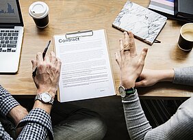 pessoas discutindo em uma mesa com computador e papel onde se lê "contrato" em inglês