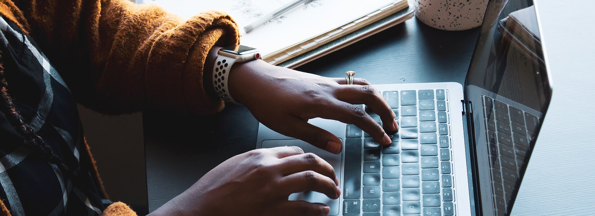 A foto mostra as mãos de uma mulher negra, que usa um relógio, mexendo em um notebook. Ao lado do notebook tem um caderno e uma xícara. 