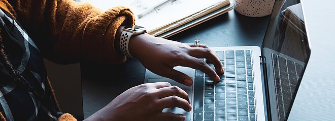 A foto mostra as mãos de uma mulher negra, que usa um relógio, mexendo em um notebook. Ao lado do notebook tem um caderno e uma xícara. 