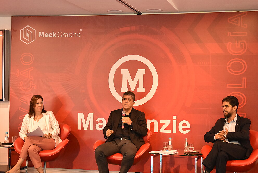 Na foto, da esquerda para direita, estão: Janette, Marco Tullio e Marcelo Coelho. Eles estão respeitando o distanciamento social. Logo atrás, tem um banner do Mackenzie, com a marca e o logo do Mack Graphe.  