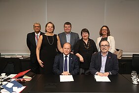 Sete pessoas posam para foto em frente à mesa de assinatura do memorando de entendimento
