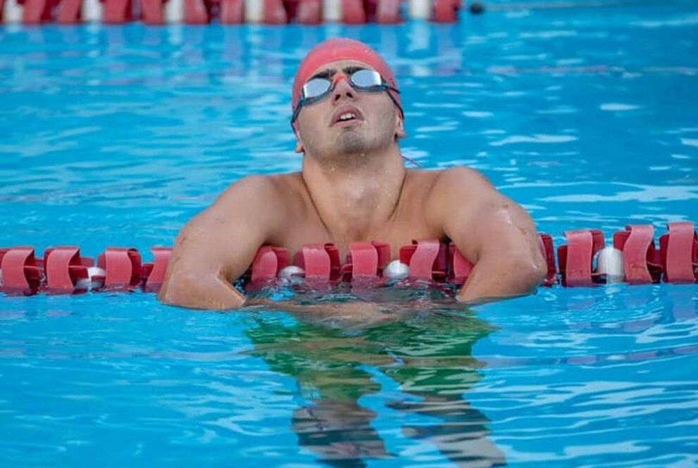 homem de óculos de natação dentro de piscina escorado nas raias