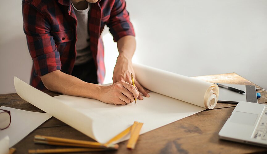 A foto mostra um homem com um lápis na mão começando a desenhar em uma cartolina em branco. Na mesa tem outras folhas e réguas espalhadas. Não conseguimos ver seu rosto.