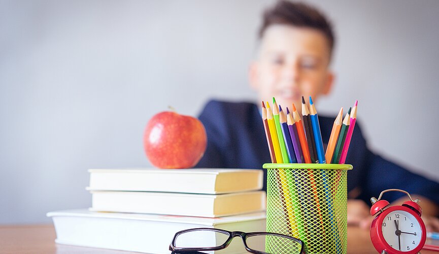Professor sentado em sua mesa com livros, canetas, relógio e uma maçã.