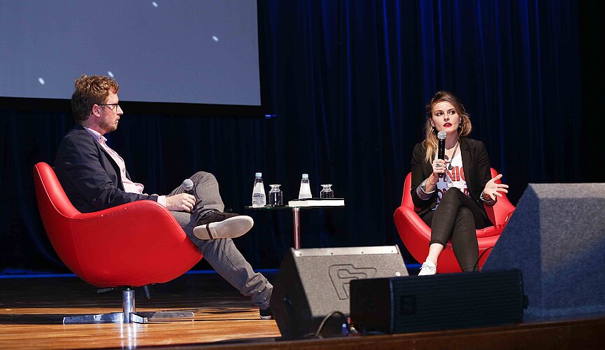 Leandro Narloch e Gloria Álvarez sentados em poltronas vermelhas sobre o palco do auditório Ruy Barbosa.